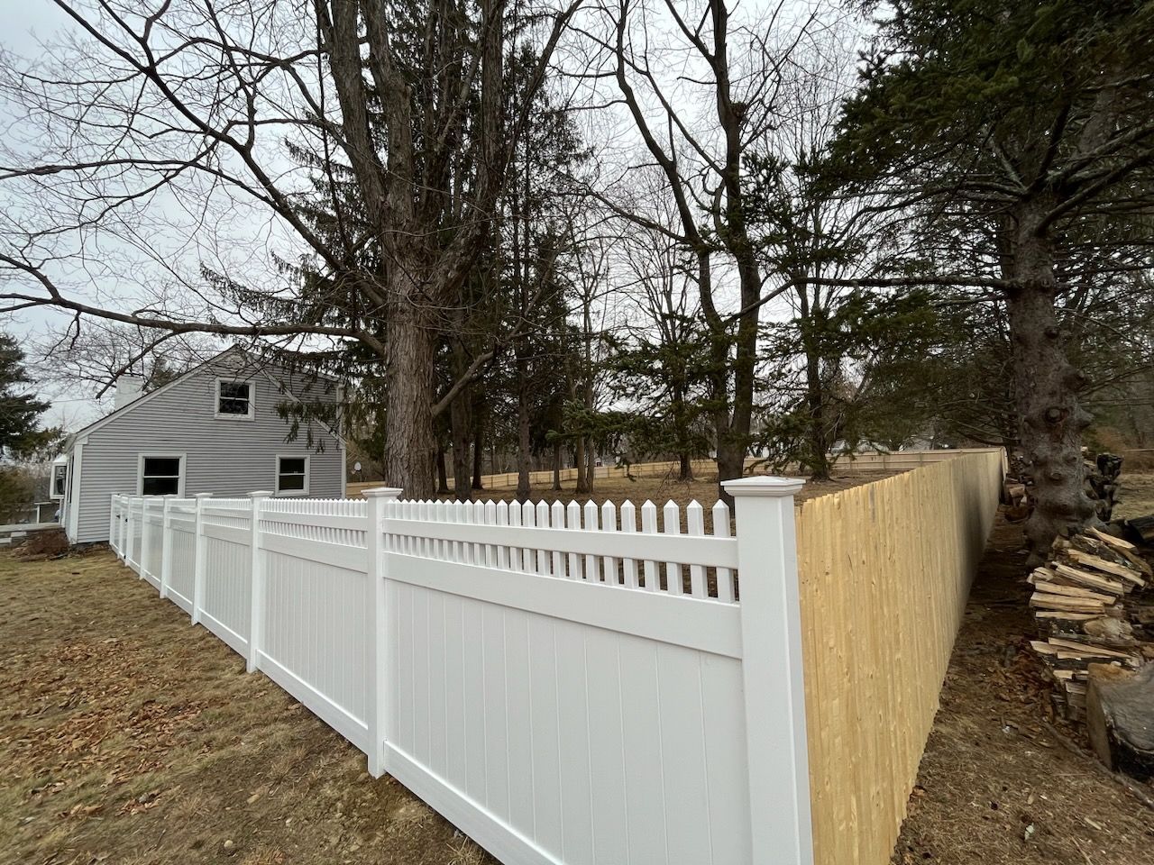 White vinyl fence beside a wooded yard and gray house under bare trees
