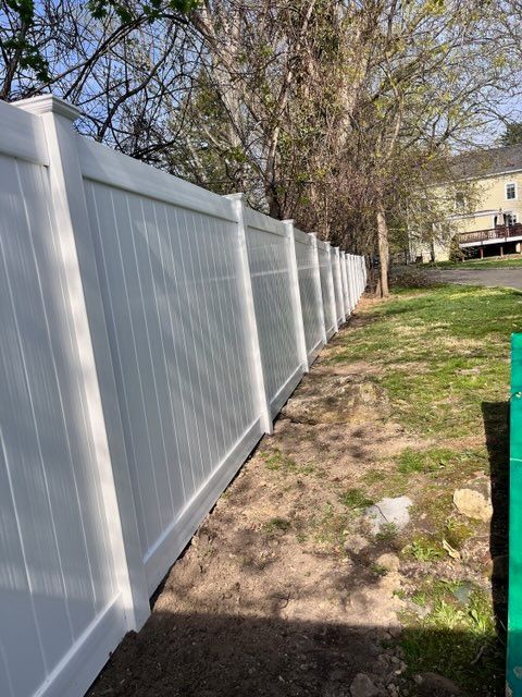 White vinyl fence beside a patchy grassy yard with trees and a house in the background