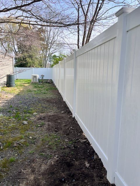 Narrow side yard with gravel and dirt beside a long white vinyl fence and bare trees