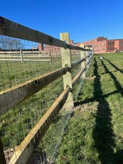 Wooden fence beside grassy field, with a brick building in the distance under a clear blue sky.