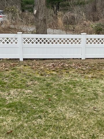 White vinyl fence with lattice top in a grassy yard, with trees and brush behind it.