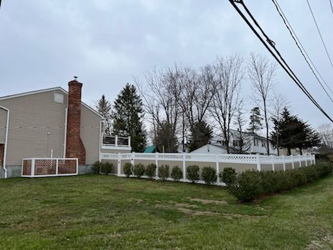 Backyard with white fence, beige house, brick chimney, and grassy lawn under a cloudy sky