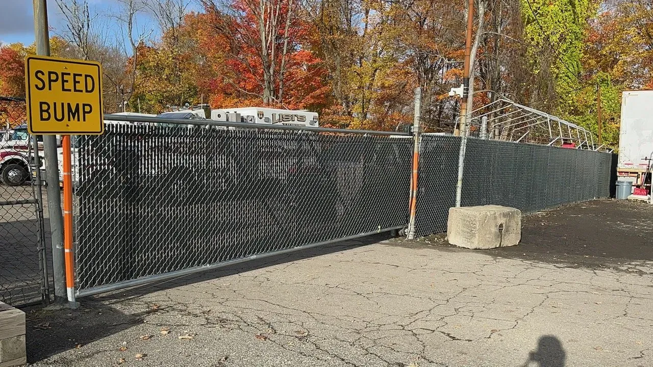 Yellow speed bump sign beside black fenced area on gravel lot with autumn trees behind
