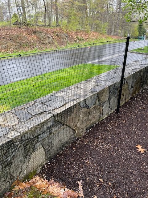 Stone retaining wall beside a wet road with wire fence and a green grass strip in a wooded area