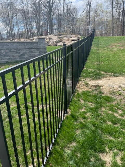 Black metal fence beside green grass and a stone retaining wall in a wooded yard