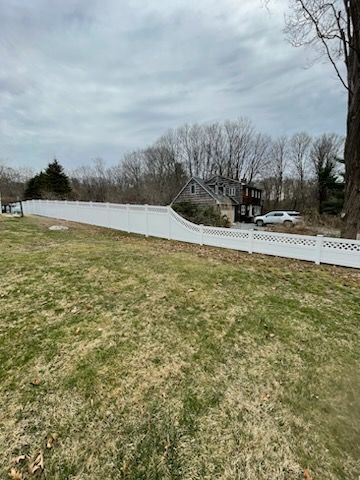 Grassy yard with a white vinyl fence, bare trees, and houses under a cloudy sky