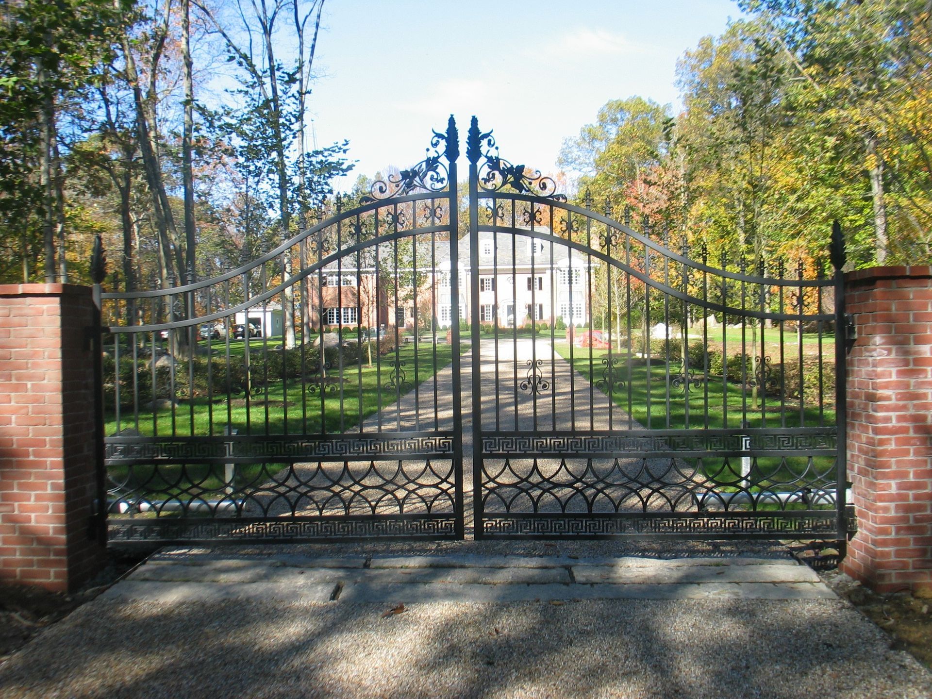 Closed black iron gate at a brick entrance with a tree-lined driveway and white house beyond
