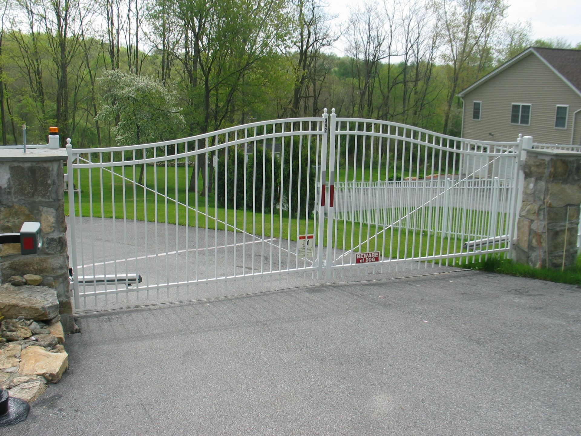 White metal driveway gate across a paved entrance beside stone pillars and trees