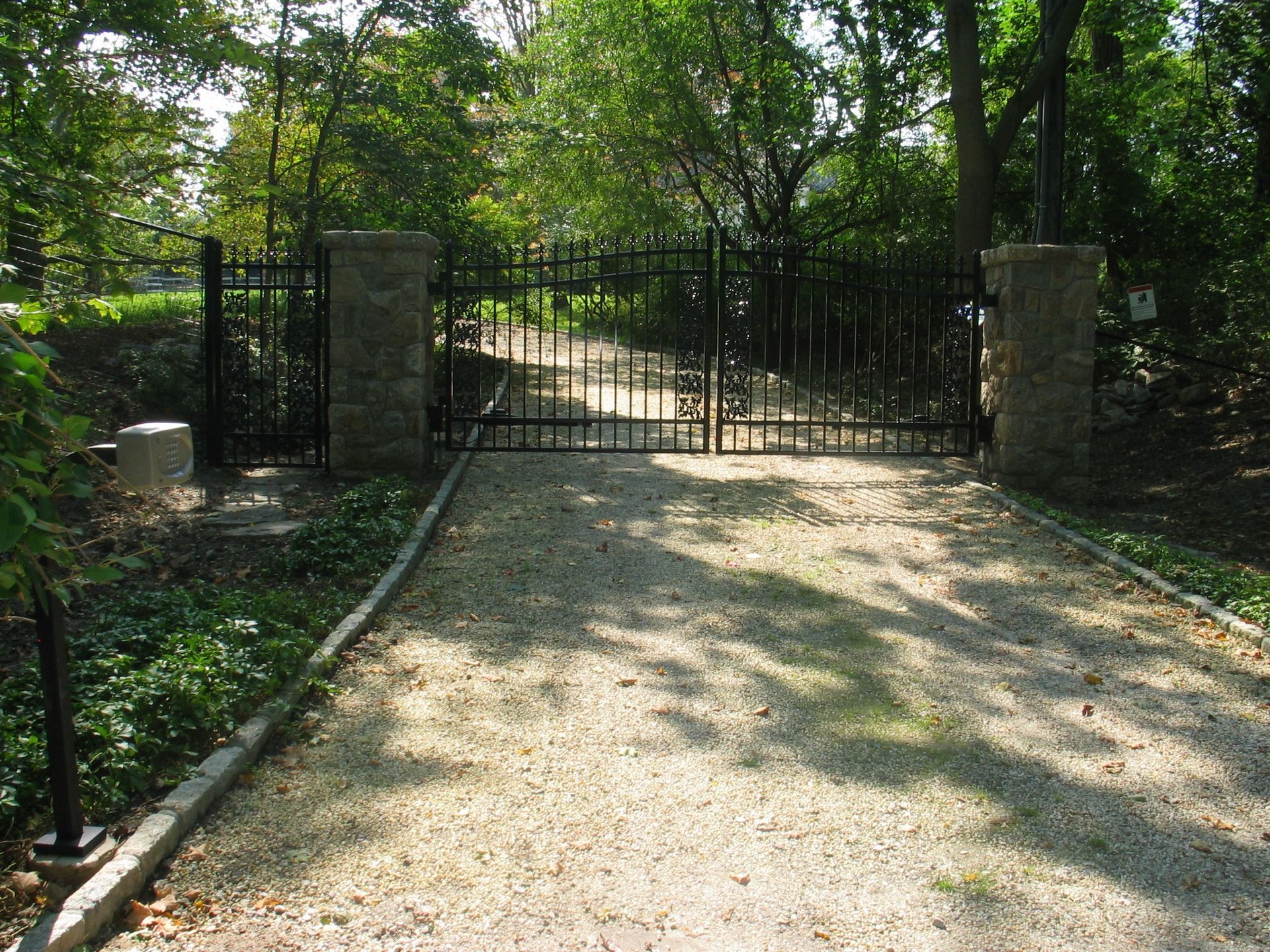 Gravel driveway leading to a black metal gate between stone pillars in a wooded area