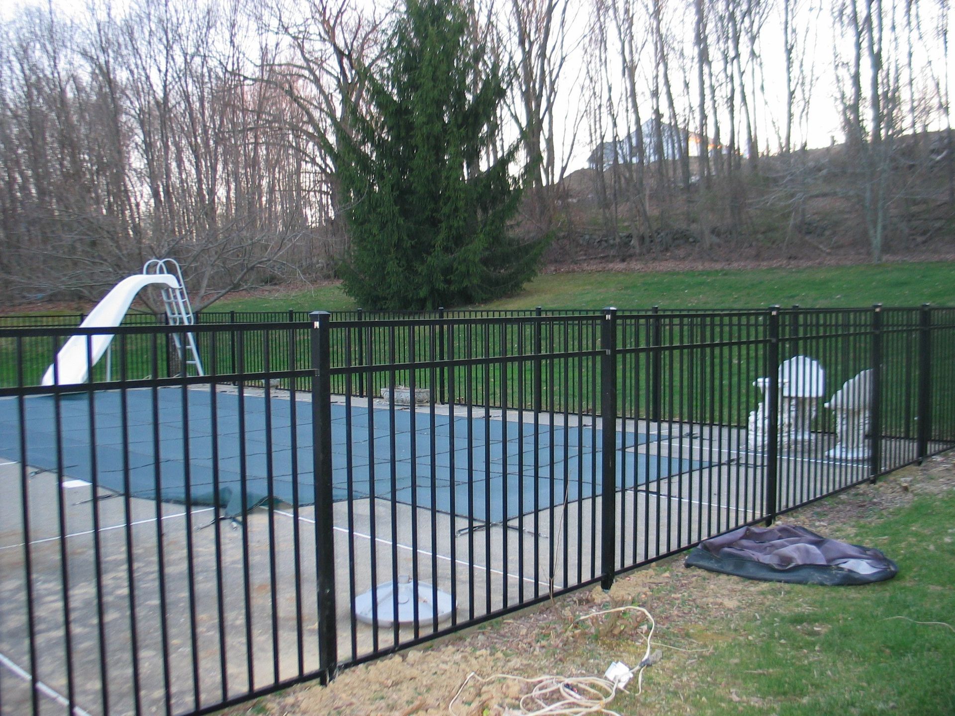 Backyard pool covered with blue tarp behind a black metal fence, with slide and lawn beyond.