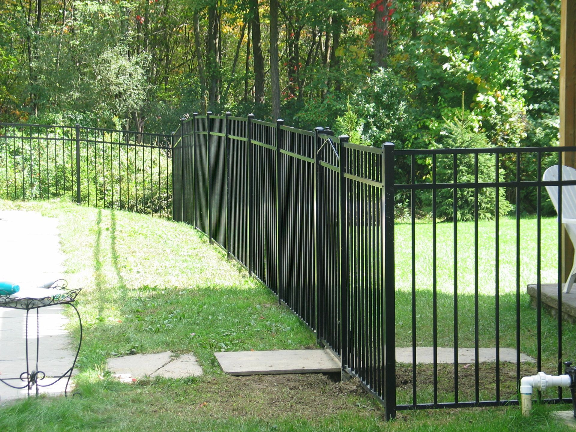 Black metal fence along a grassy path in a sunny backyard with trees and stepping stones