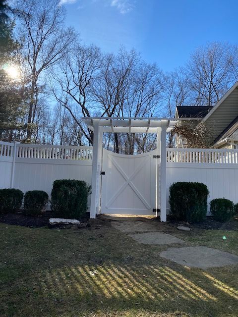 White backyard gate with trellis, surrounded by fence and trimmed shrubs under a blue sky.