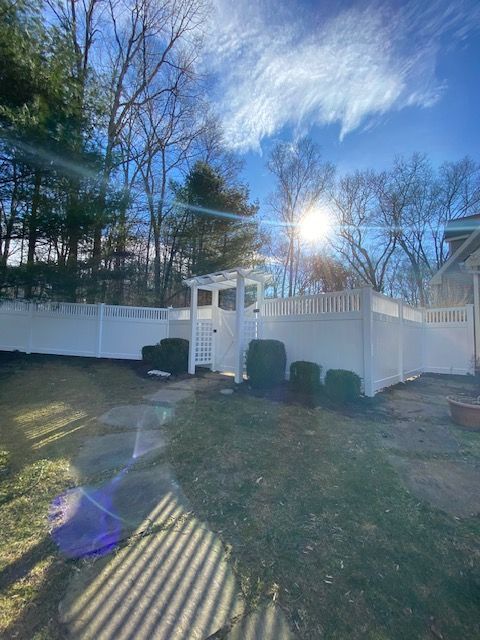 Backyard with white fence and gate, bare trees, bright sun, and a lens flare over the lawn.
