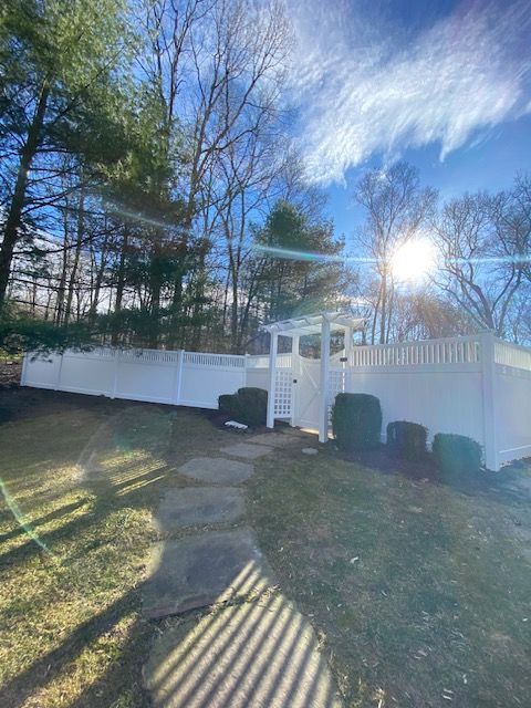 Backyard with white fence, stone path, small gate, and bright sun through tall trees
