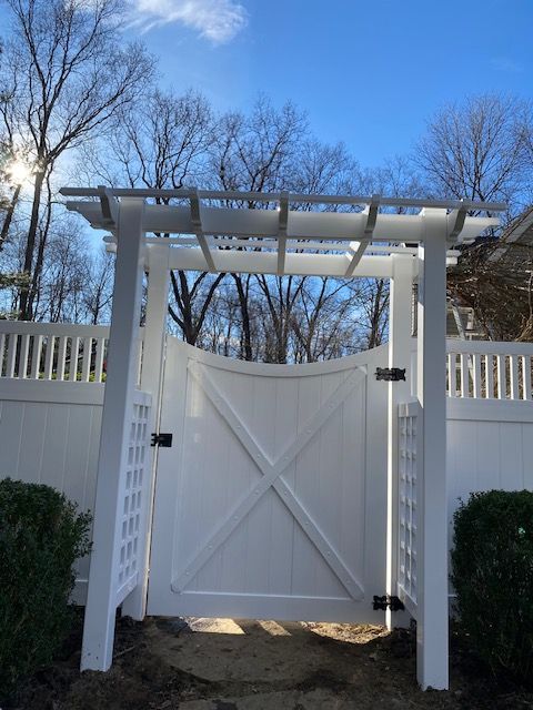 White garden gate with pergola, flanked by white fencing and evergreens under a bright blue sky.