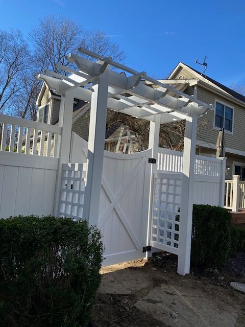 White pergola gate in a fenced backyard beside a beige house, with bushes and a blue sky.