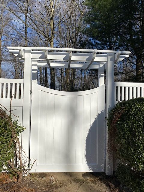 White garden gate with pergola-style top between matching fences, in front of bare trees and blue sky.