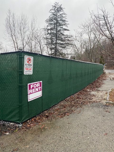 Green construction fence with a “For Rent” sign along a gravel roadside in winter.
