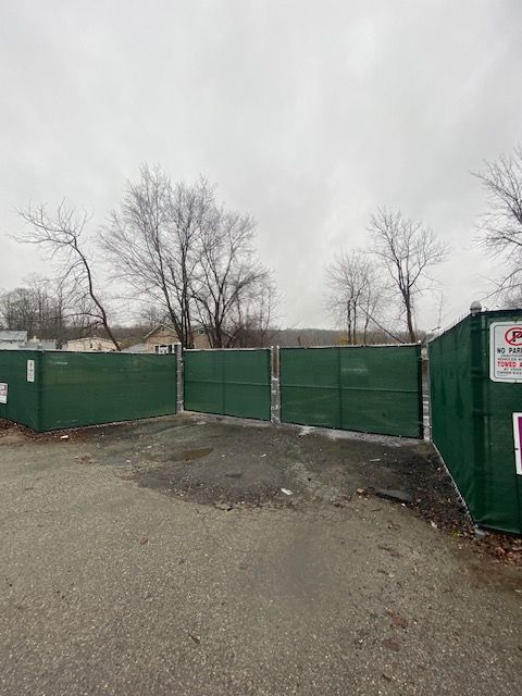 Green construction fence blocking a gravel lot under a gray sky, with bare trees behind it.