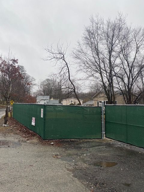 Green construction fence beside a wet roadside with leafless trees and overcast sky