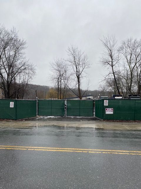 Green construction fence blocking a wet roadside, with bare trees under a gray sky