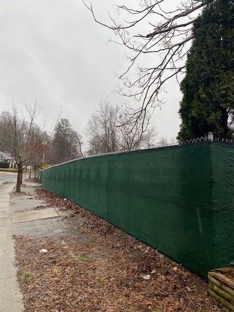 Rainy sidewalk beside a long green construction fence under bare trees