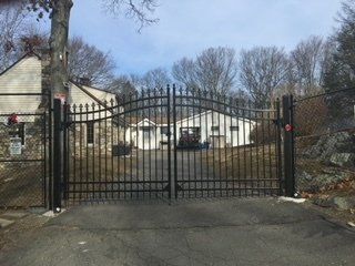 Black iron gate blocking a driveway in front of a house, with trees and a stone wall nearby.