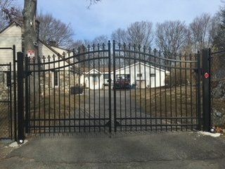 Black wrought-iron gate blocking a driveway in front of a white house and leafless trees