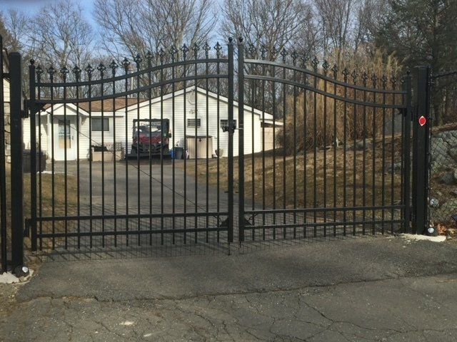Black wrought-iron driveway gate in front of a white house with a parked vehicle behind it.