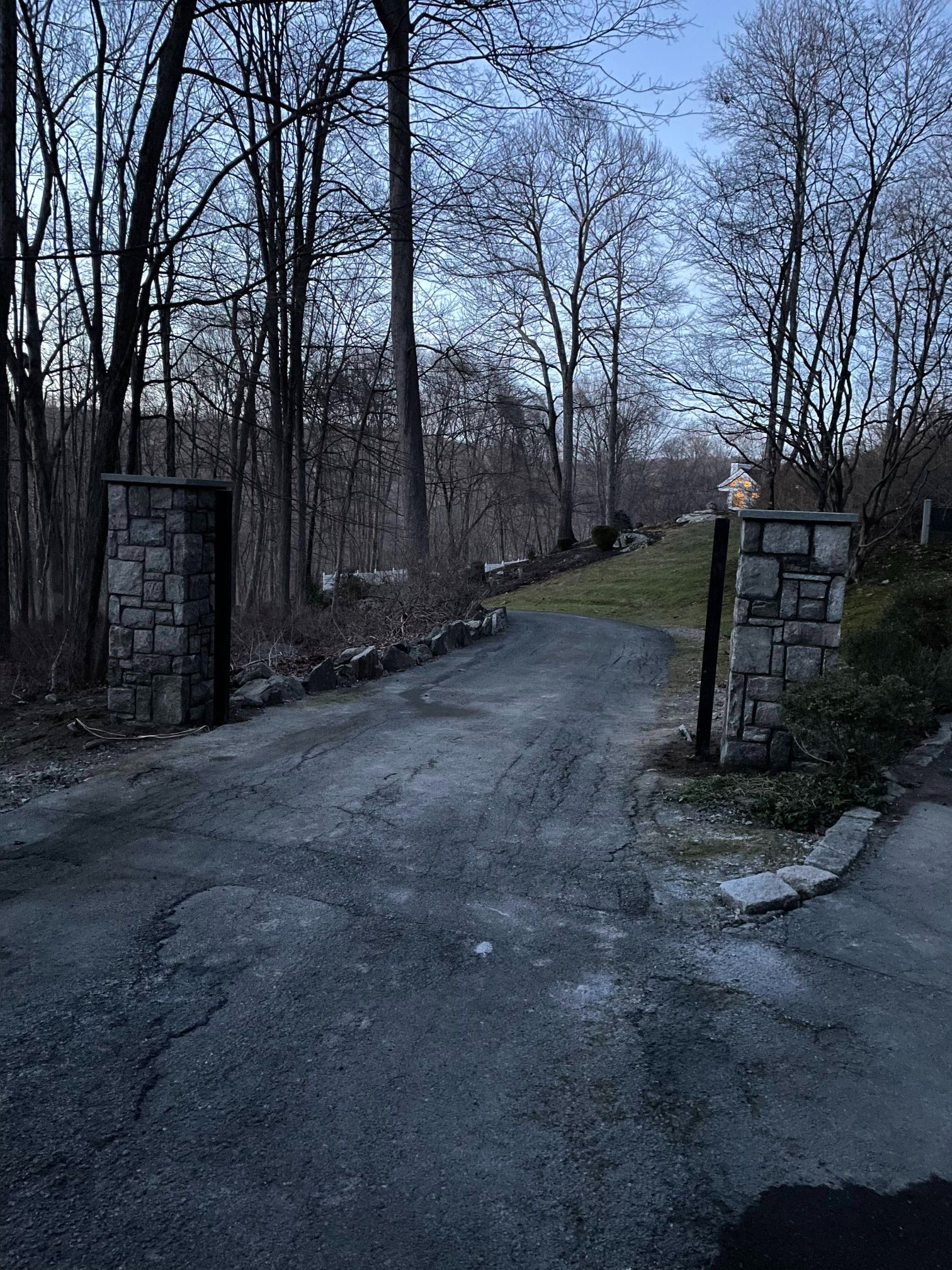 Stone-pillared driveway curves uphill through bare trees at dusk.