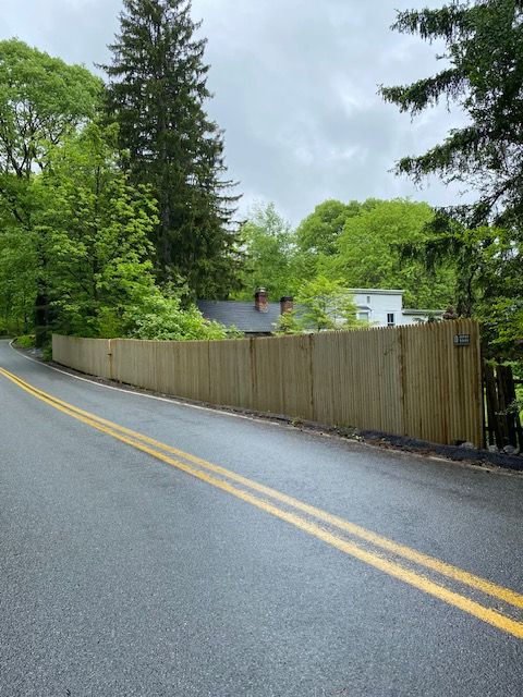Wet two-lane road beside a wooden fence and green trees on a cloudy day