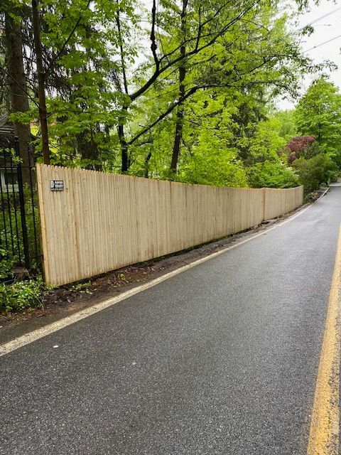 Wet paved road beside a beige wooden fence and green trees on a cloudy day
