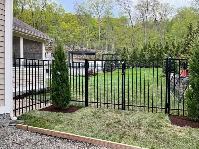 Black metal fence enclosing a small yard beside a house, with shrubs and trees in the background.