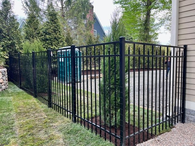 Black metal fence around a small gravel yard beside a house, with trees and a blue utility box in the background