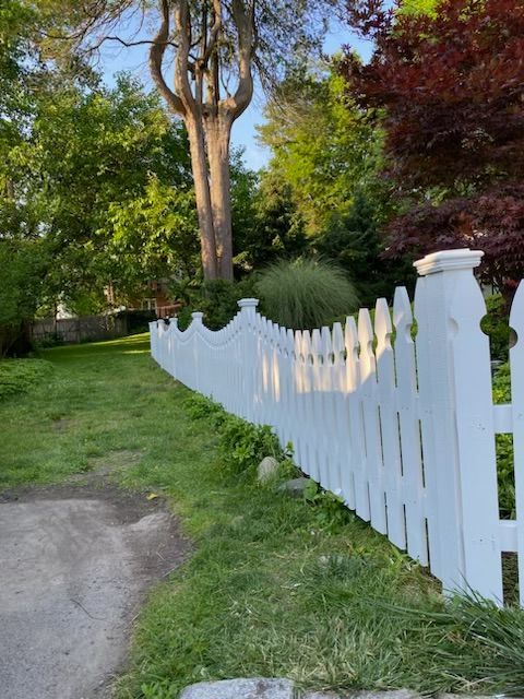 White picket fence along a grassy path in a green garden with tall trees.