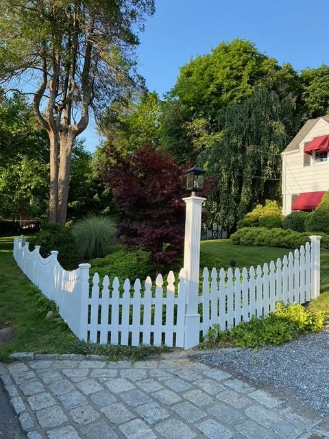 White picket fence around a small garden beside a cobblestone path, with trees and a house in the background.