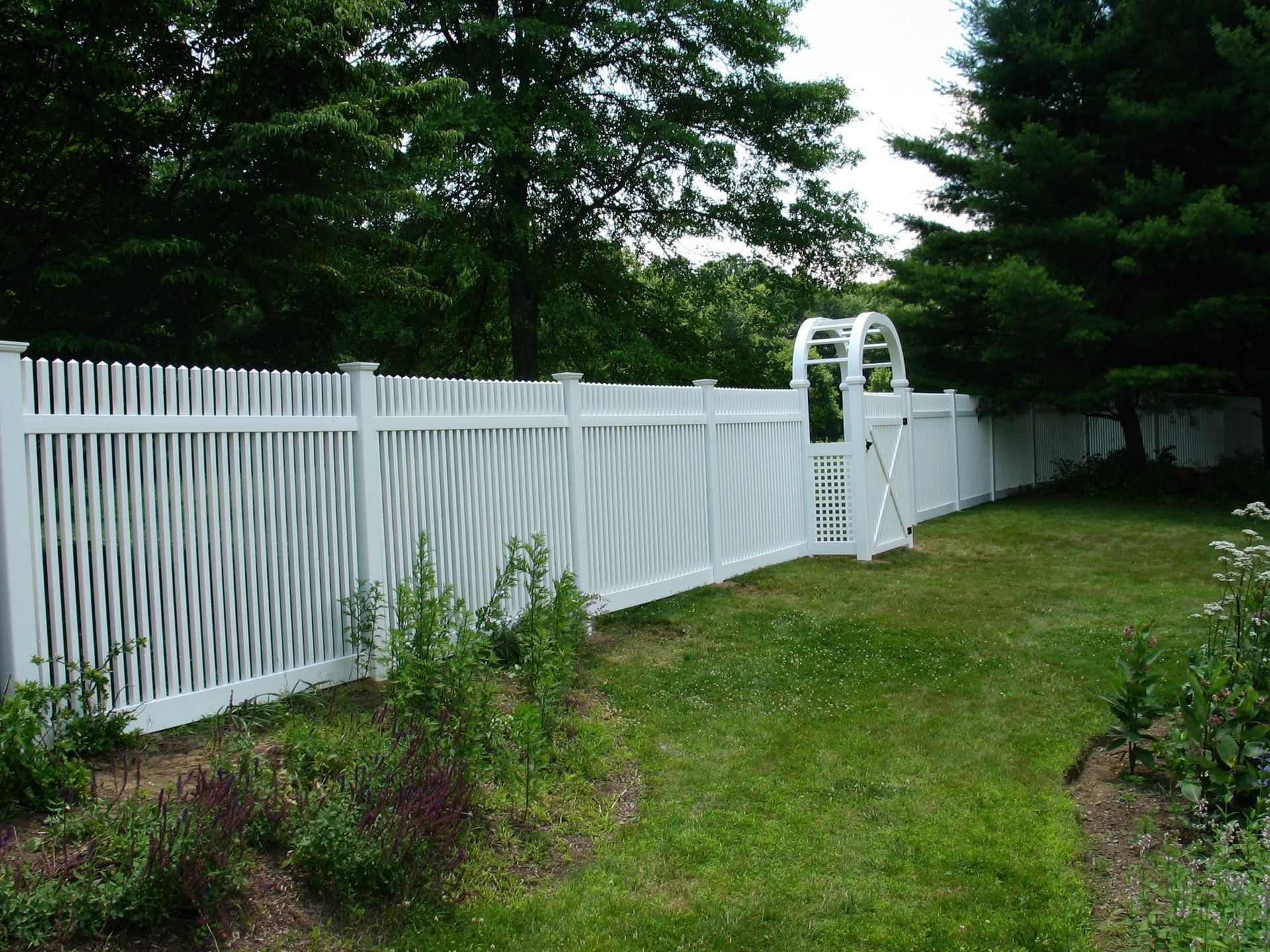 White picket fence with an arched gate along a grassy backyard bordered by trees and shrubs