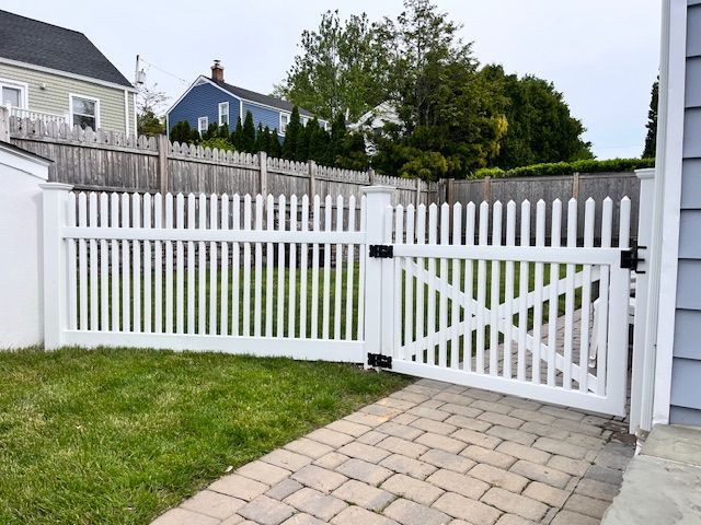 White wooden fence with a gate beside a paved walkway and green lawn in a residential yard.