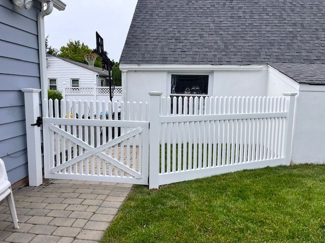 White picket fence and gate beside a gray house, with a paved walkway and green lawn.
