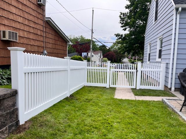 White vinyl fence and gate enclosing a small grassy backyard beside a house with a narrow walkway.