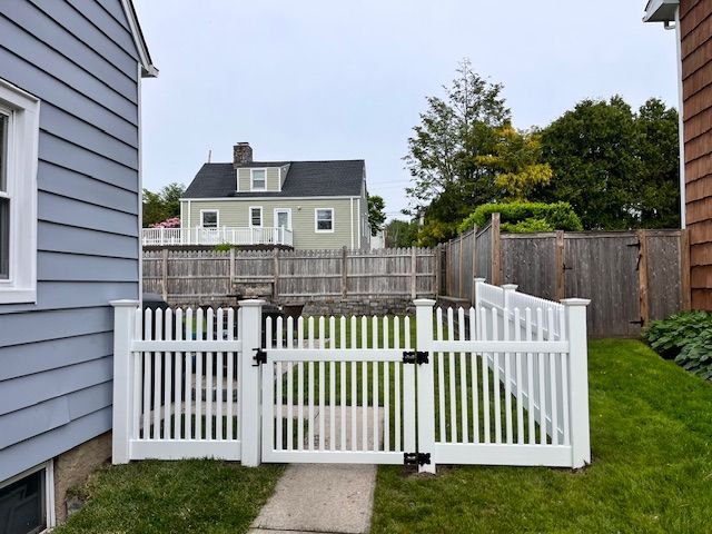 White picket fence and gate enclosing a small backyard beside a gray house, with a lawn and neighboring homes.