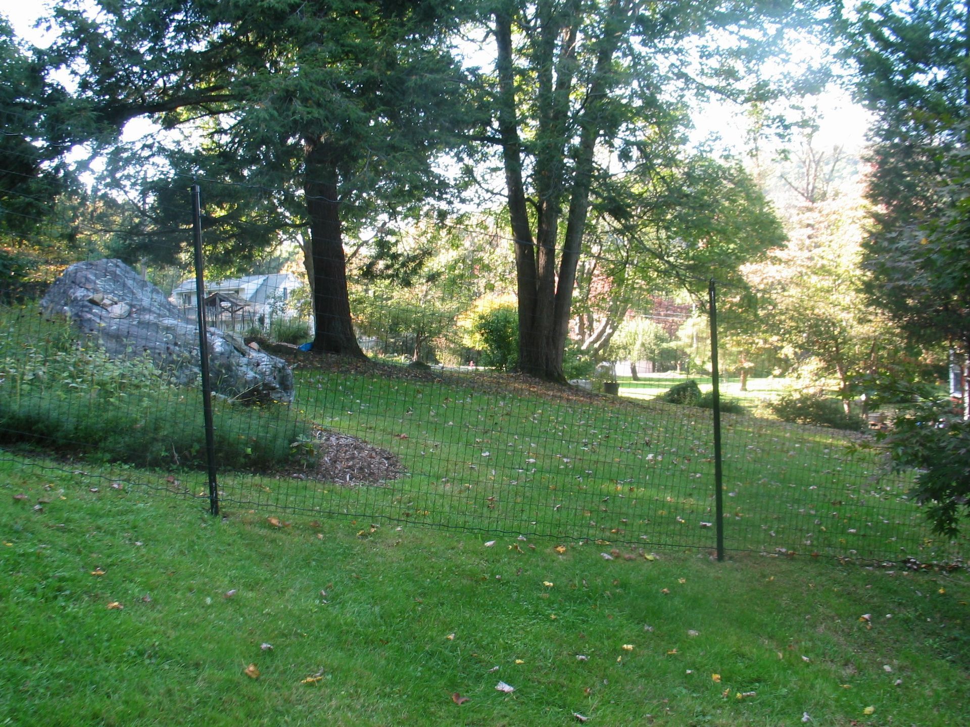 Shady park lawn with trees, a large rock, and two black posts under dappled sunlight