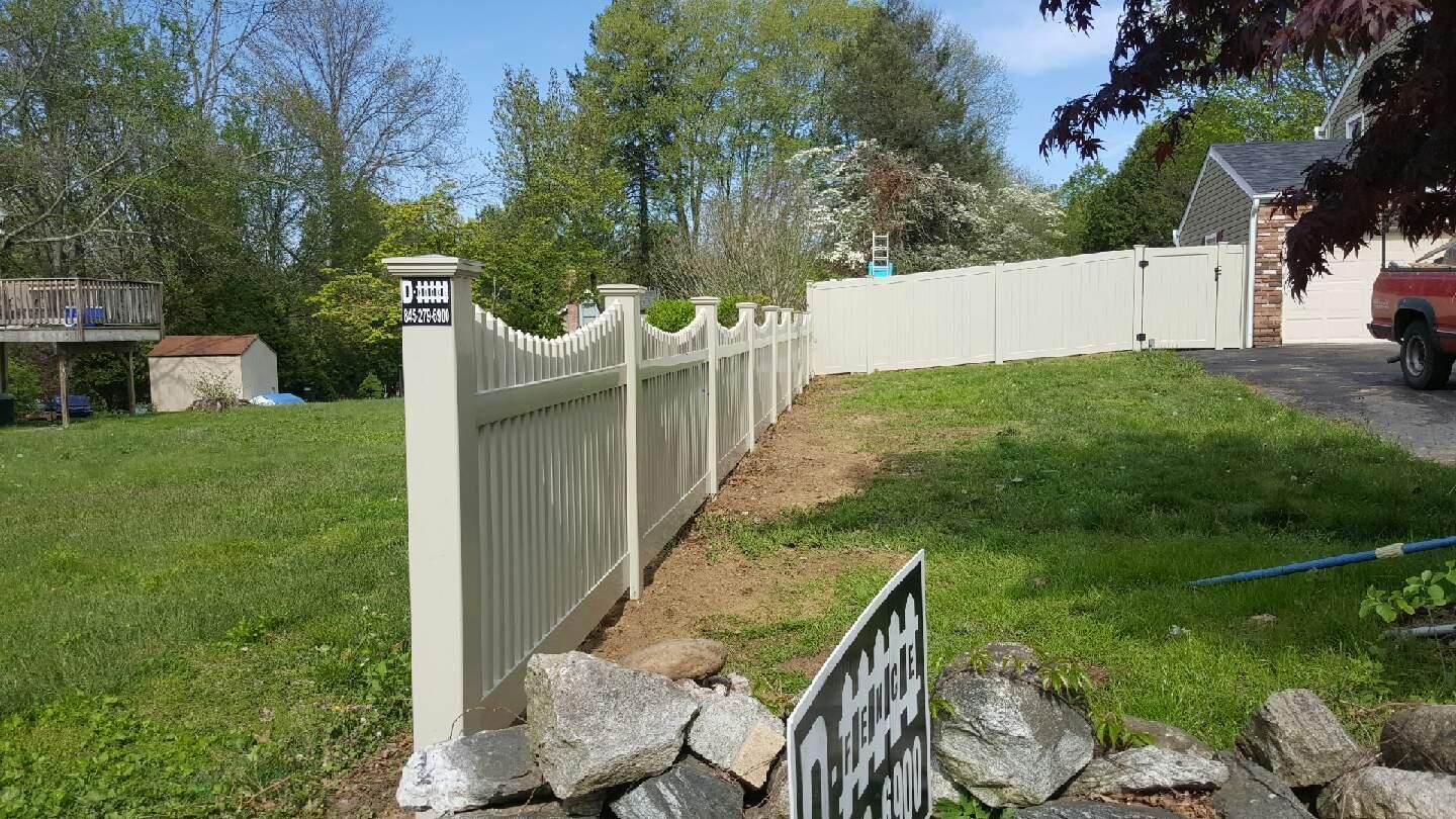 White vinyl fence along a grassy yard with rocks in the foreground under a clear blue sky.