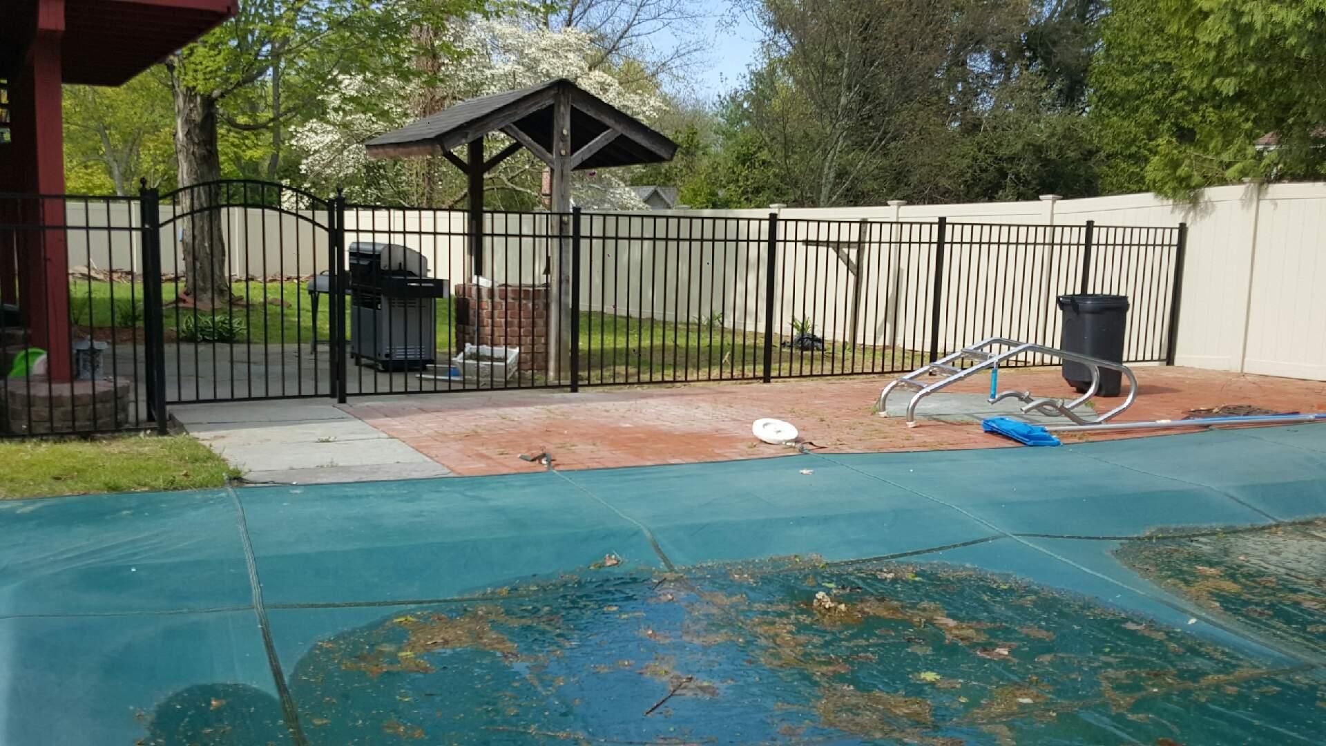 Backyard pool area with a covered pool, fence, patio, and scattered cleaning tools under trees.