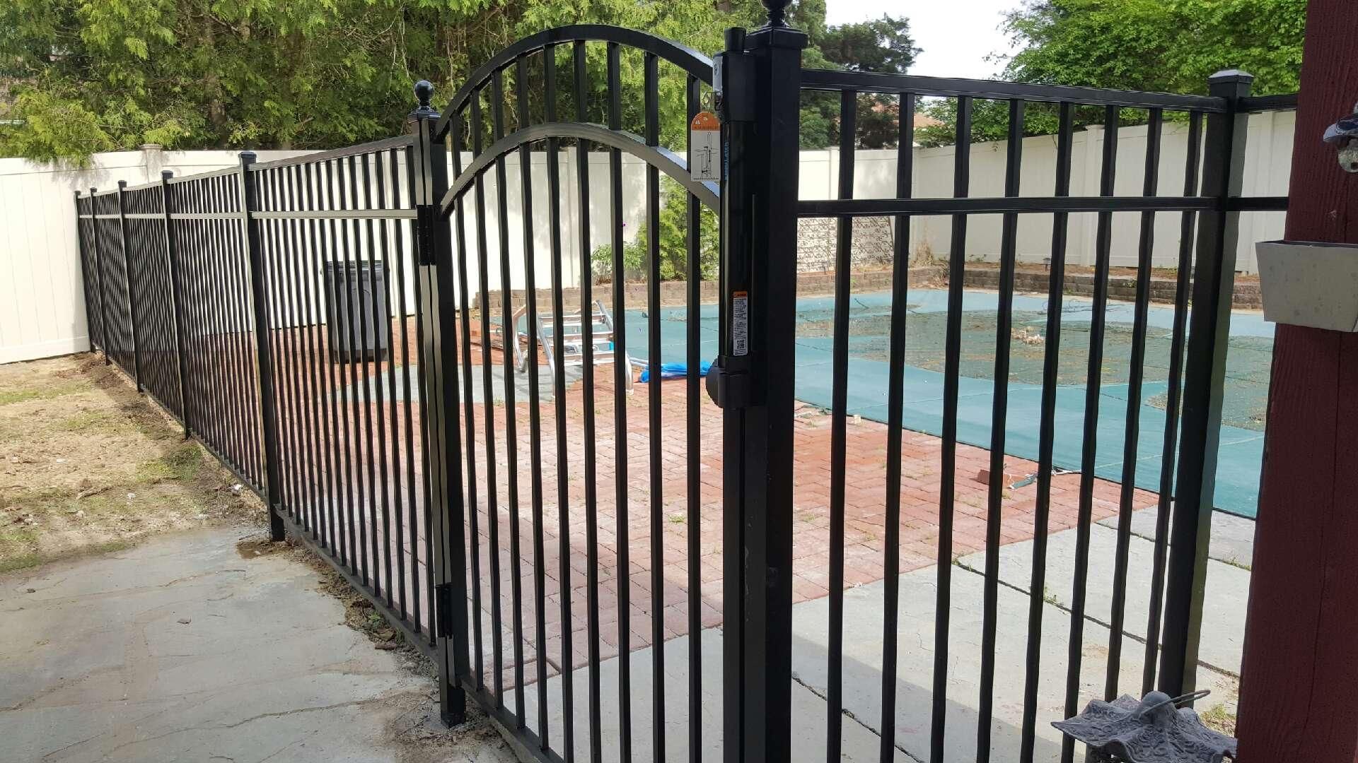 Black metal fence beside a backyard swimming pool with brick patio and trees in the background