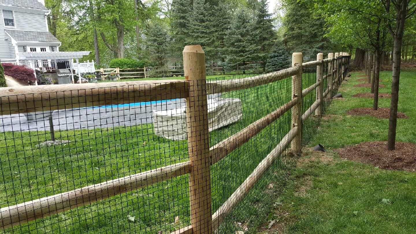 Wooden split-rail fence along a grassy yard beside a road and trees.