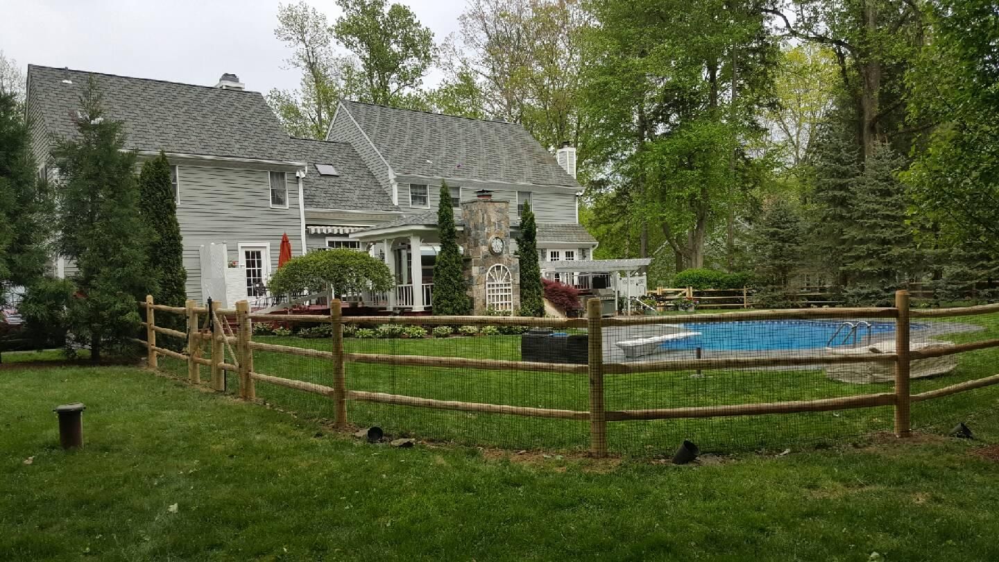 White house with gray roof beside a fenced backyard and blue pool, surrounded by trees and grass.