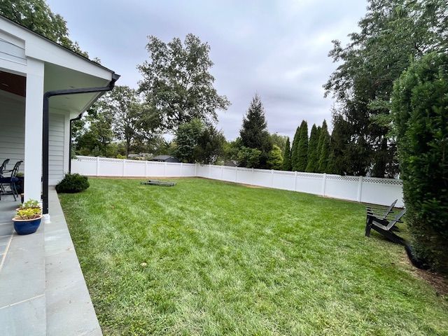 Backyard lawn with white fence, trees, patio, and potted flowers beside a house