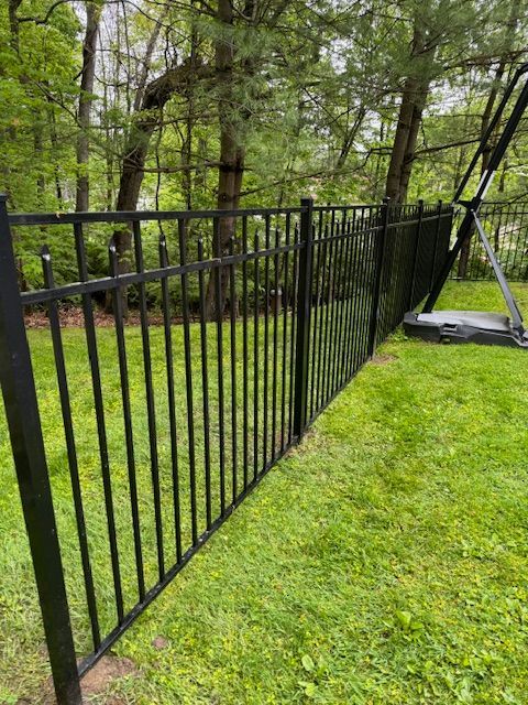 Black metal fence beside a green lawn and wooded area, with a leaning gate section.