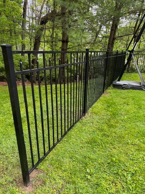 Black metal fence beside a green lawn and wooded area, with a sloped panel near a patio.